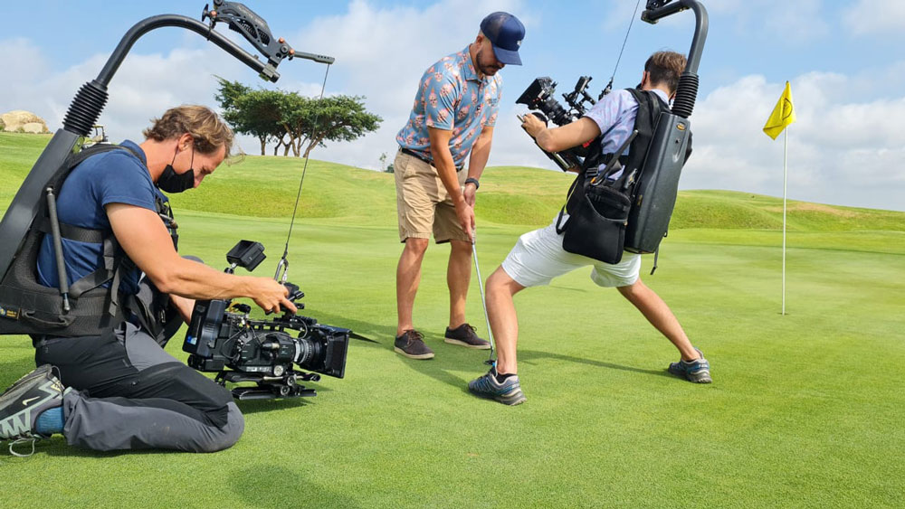 Two men wearing equipment and holding video cameras taking videos of a man holding a golf club and getting ready to hit it.
