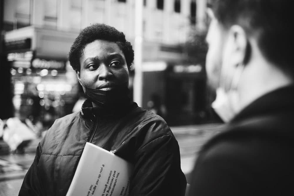Person with their mask lowered and holding pieces of paper. They are looking at another person.