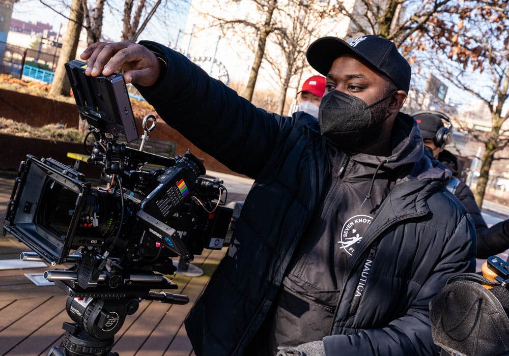 Man wearing a black jacket, hat, and mask and looking into a video camera.
